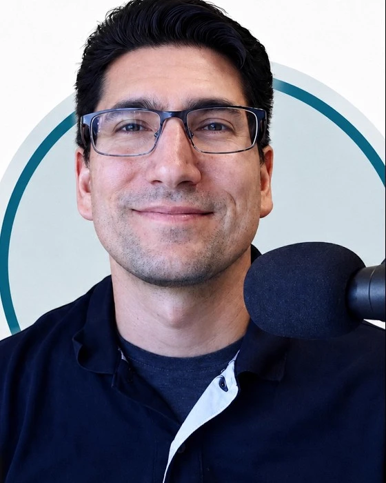 Doug Spence, smiling, wearing a navy polo and glasses, at his podcast microphone.